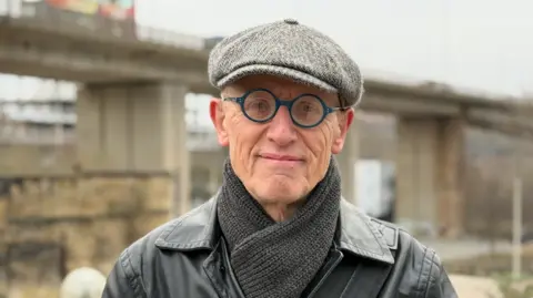 BBC A man wearing a grey cap, a grey scarf and circular blue rimmed glasses stands in front of a beige-coloured concrete bridge.