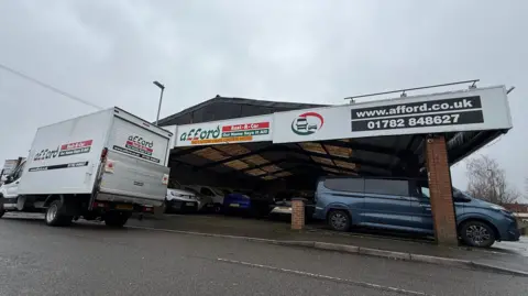 A garage with vehicles parked under a large roof. A white van is parked at the side of the road in front of it.