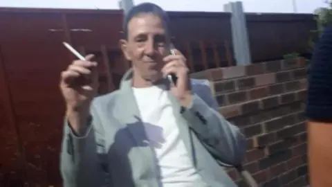 David Aubert, in his 50s, who has dark blonde hair, sits on a chair in a back yard holding a cigarette in his right hand and a mobile phone to his ear in his left hand. He is wearing a light grey blaze over a white t-shirt.