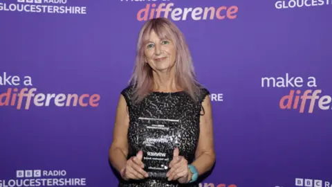 A woman with shoulder length pinky blonde hair smiles as she holds a clear, square-shaped award. She is wearing a black, sleeveless sequin dress and is standing in front of a purple background with BBC Radio Gloucestershire Make a Difference branding on it.