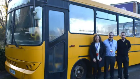St Augustines Academy A bright yellow bus parked on the school playground, with three people standing in front. They are, from left to right, Amanda Howes, Steve Mead and Jason Smart. They are all smiling at the camera.