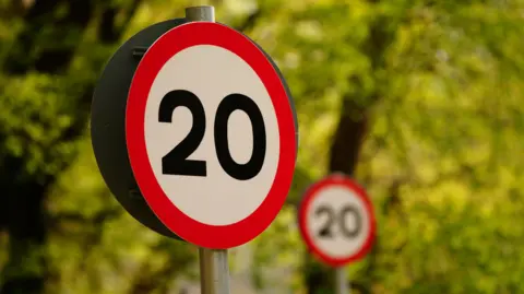 A 20mph sign in the fore with another out of focus behind it, with a backdrop of green trees.