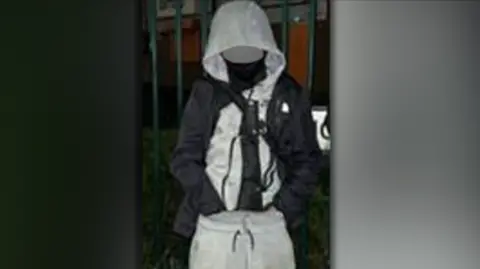 West Midlands Police A boy in a gray tracksuit, with a machete in a black sheath in his trousers. His face is blurry and he is standing outside in the dark in front of a fence