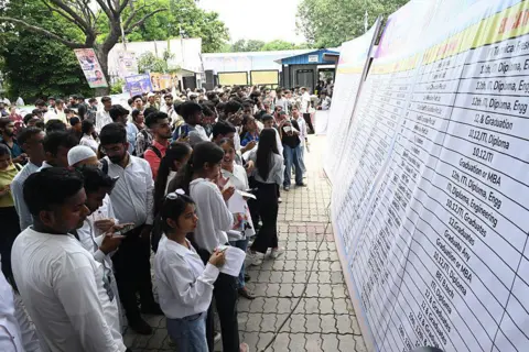 Hindustan Times via Getty Image Dozens of young people, men and women, stand in front of a big board listing out educational qualifications for different jobs