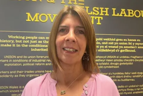 Rosie Lewis smiles at the camera. She has shoulder-length straight auburn hair and earrings in the shape of a star. She is wearing a pink shirt.