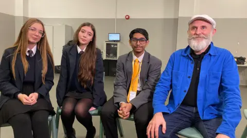 Two girls and a boy in school uniform sitting next to a bearded man wearing a cap and bright blue jacket