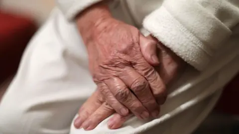 Close up on: An old person in a dressing gown with papery skin clenches one hand with the other.