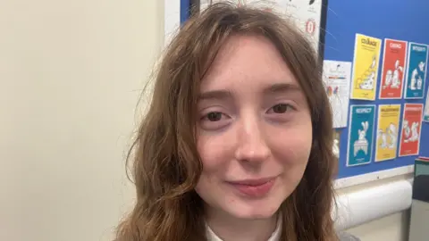 Polly is smiling at the camera. She has curly brown hair and is wearing a white shirt and grey blazer. In the background a blue notice board displays yellow and red pages with motivational quotes.