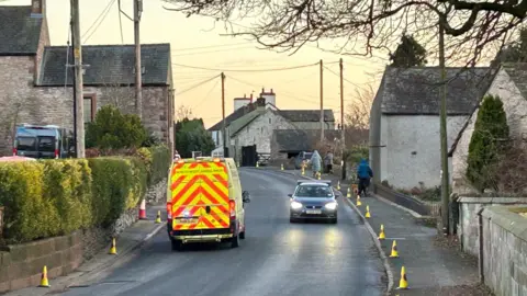 An ambulance is driving on a two-lane road in Clifton. A car is travelling in the opposite direction. There are stone houses on each side of the road. Three people are walking on the pavement. 