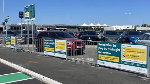 Signs at the airport drop-off attached to a silver fence that remind people to pay online or by phone by midnight the next day.