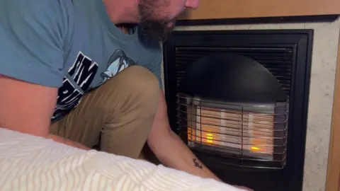 A man lighting a gas fire in a caravan with the bed in the foreground