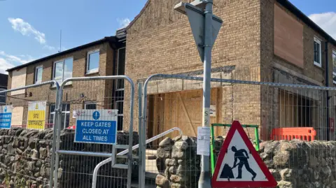 View of a boarded up two-storey building, There is a metal fence surrounding the site and a "men at work" road sign.