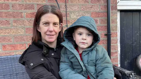 Kate Bradbrook/BBC A woman in a black coat and a boy in a green coat pose in front of a brick wall.