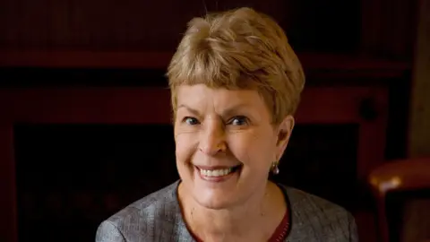 Getty Images A woman with short blonde hair smiles at the camera. She has pearl drop earrings and wears a grey jacket with a red top underneath. 