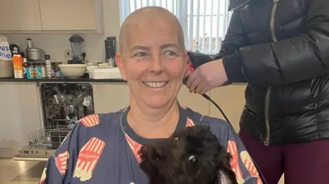 Family photo Lisa Noble smiling in a kitchen while having her head shaved during cancer treatment. She is wearing a blue T-shirt with illustrations of buckets of popcorn on it. A small black dog is sitting on her lap and has quickly turned to the camera with its eyes wide, creating an amusing scene. There is a woman with a black puffer jacket and maroon leggings holding an electric shaver next to her. Her face is off-camera.