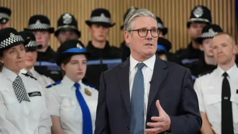 PA Media Sir Keir Starmer is wearing a dark blue suit jacket, on top of a white shirt with blue tie. He's giving a speech with several male and female police officers standing behind him.