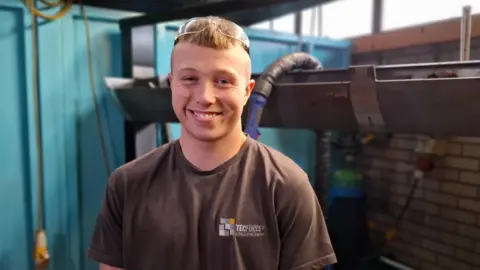 Conner is wearing a Tecforce T-shirt with protective goggles resting on his head. He is stood in the training facility smiling at the camera. Conner has blonde hair and no facial hair.