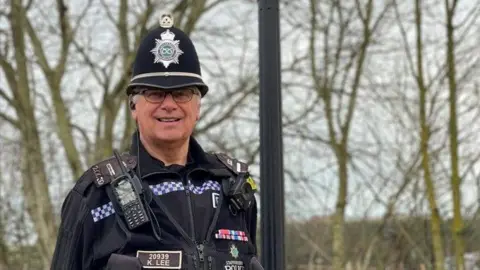 Staffordshire Police Police officer Kevin Lee is stood next to a lamppost. He is wearing glasses and black police uniform with a body-worn camera, police radio and other police equipment attached. There are trees in the background.