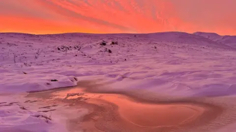 the sun rising over the Cairngorm plateau has turned the sky and the snowy ground a deep pink colour. There is an icy pool in the foregound.