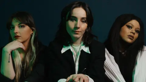 Sarah Ward Cherym, made up   of L-R Allanagh Doherty, Emer McLaughlin and Hannah Richardson. The set  are dressed successful  monochrome and beryllium   posing connected  a achromatic  sofa against a acheronian  bluish  backdrop. 