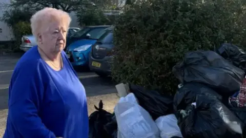 An older woman with pink hair and wearing a blue top stands next to a pile of bin bags outside a car park. The pile is nearly a tall as she is.