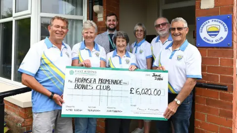 A group of three women and four men stood outside Hornsea Promenade Bowls Club. Three women and three men are wearing blue, white and yellow bowls kit. The other man is wearing a striped, blue shirt and a tie. Some of them are holding a large check for £6,020.00 written to Hornsea Promenade Bowls Club.