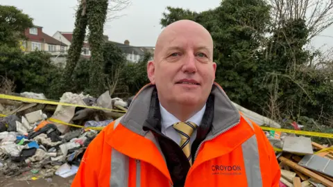A man in orange hi viz looks into camera, he is bald with a small smile, yellow tie and on the high Viz it says Croydon 