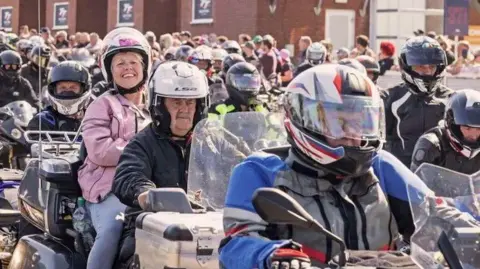 IOM TT A large group of motorcyclists in biker gear lined up in the pit lane at the grandstand. A lady wearing a pink jacket on the back of a bike smiles broadly.
