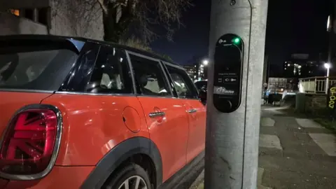 A black charging point is stuck to a lamp post with a green light, next to a red mini. 