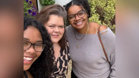 Family photograph Caroline poses for a family photo with her daughters Francesca and Eva. Francesca is wearing glasses and smiling. She has curly black hair. Eva also has glasses and has black hair tied back with gold jewelry and a stone coloured jumper. Caroline is in the centre with a brown and black dress.