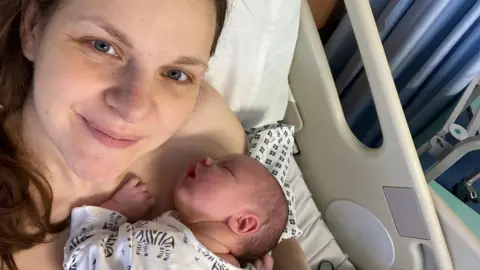 Deborah Pearson A selfie of a woman in a hospital bed, holding her newborn baby who is asleep, she smiles towards the camera 