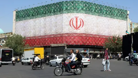 Iranians drive cars and motorbikes past a billboard featuring Iran's national flag at Enqelab Square in Tehran, Iran.