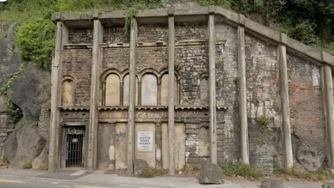 The remaining façade of the Clifton Rocks Railway. Its signage is barely visible underneath the concrete pillars installed to keep the structure from collapsing. Its arched tunnels and windows have been filled in and there is a rusty gate blocking the door.