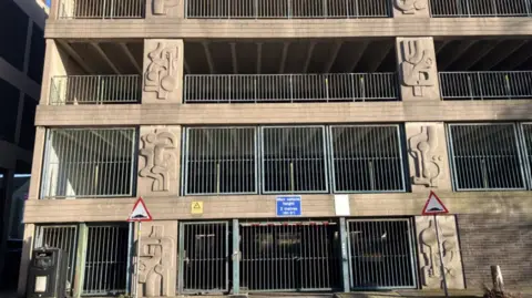 Lara King/BBC A tan coloured multi-storey car park which is made of concrete with concrete sculptures on the building. The art work is abstract. There are railings and a gate which is closed. It is described as a 'brutalist' building. 