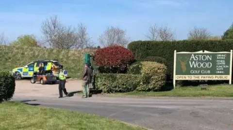 The entrance of a golf course, with police setting up a cordon outside of it. Green bushes line the entrance with a sign saying "Aston Wood Golf Club".