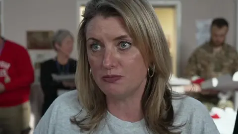 Louise Fetigan looking at the camera with a serious expression, she has blonde hair, dark lipstick on, hoop earrings and a white top. In the background are people dressed in military uniform and jumpers
