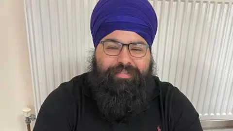 Kuljit Singh Uppal dressed in a black top and blue turban and black glasses. He is sitting on the floor in front of a radiator 