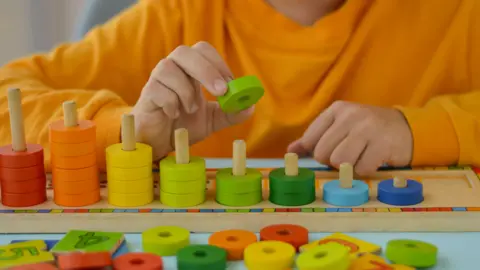 A child is wearing an orange top and is playing with a wooden maths game. The game consists of wooden pegs and coloured pieces to help count. 