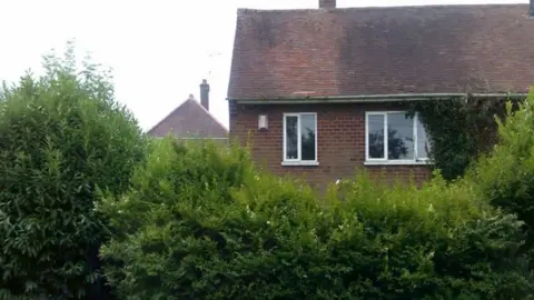 The outside of one floor and the roof are visible. Bushes are covering other parts of the house in this image taken outside. Part of another property is visible in the background.