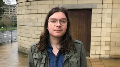 Sam Read/BBC Louis McKechnie with long dark hair wearing a green jacket and blue shirt, standing in front of a stone-built court building