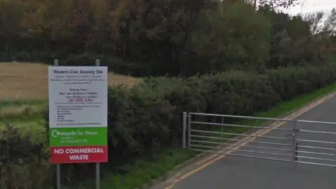Google The outside gate to the site with a sign outlining opening times that has No Commercial Waste in large letting on it.