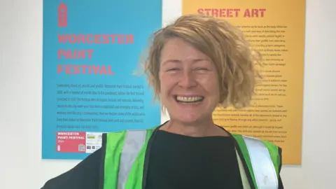 BBC Woman in a green hivis jack and black hoodie is smiling with a blue board that says "Worcester paint festival" and a yellow board that says "street art" both in bold pink writing