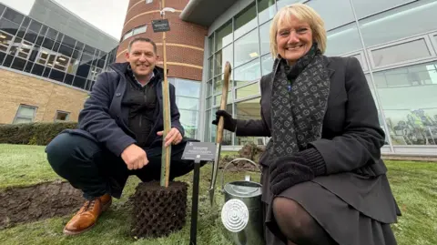 Jim Scott/BBC Damon Kent and Marion Dickson crouched down and looking into the camera with big smiles. Mr Kent is on the left and is holding the sapling in his hand with is a long, thin stick standing in a chunk of soil. He has short, brown hair and is wearing a blue coat, dark trousers and brown shoes. Ms Dickson is holding a shovel in her hand and has blonde short hair. She is wearing all black and has gloves on. The grass has been dug up on the left of them and turned up grass can be seen behind them. They are in front of the hospital which is a glass-fronted, large brick building.
