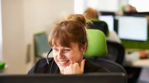 Woman wearing a headset and smiling. She has red hair with a fringe and wearing a black top. Other people at screens are in the background.