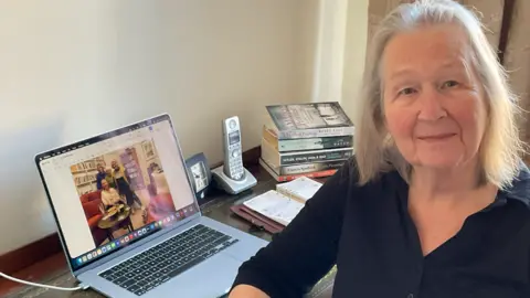 Gillian Edwards Gillian is sitting at a desk smiling to camera with a computer open beside her with a family photograph visible on the screen. She has shoulder-length grey hair and is wearing a black blouse. There is a pile of novels on the far edge of the desk and a landline phone