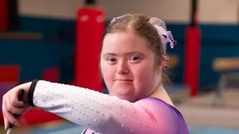 A young woman in a gymnastic training area. She is looking at the camera as she twirls a stick with a ribbon attached to it. She wears a purple and pink leotard. She has a matching coloured scrunchie in her brown hair