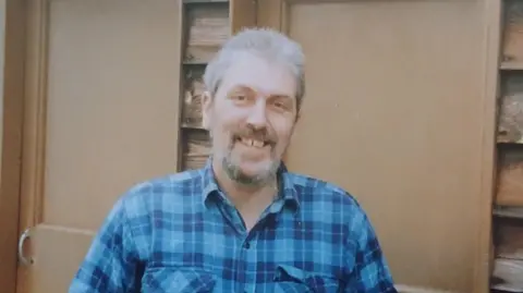 Isobel Saunders A man with grey hair and beard. He is smiling broadly at the camera is wearing a blue checked shirt.