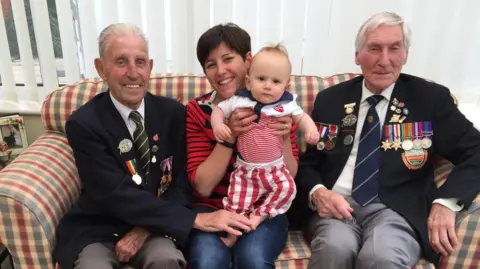 Four people are on a checkered sofa. On the left is a man wearing military medals, in the middle is a woman holding a small child, on the right is a second man wearing military medals. All are smiling.