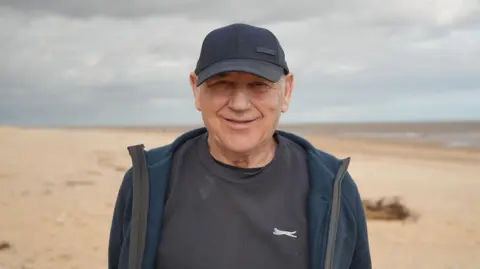 Shaun Whitmore/BBC Kenny Chaney stands on a sandy beach. he wears a navy baseball cap and dark fleece.