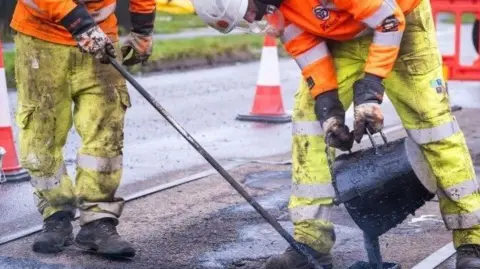 West Northamptonshire Council West Northamptonshire Council highways workers in high-viz jackets pour tarmac onto a road surface.